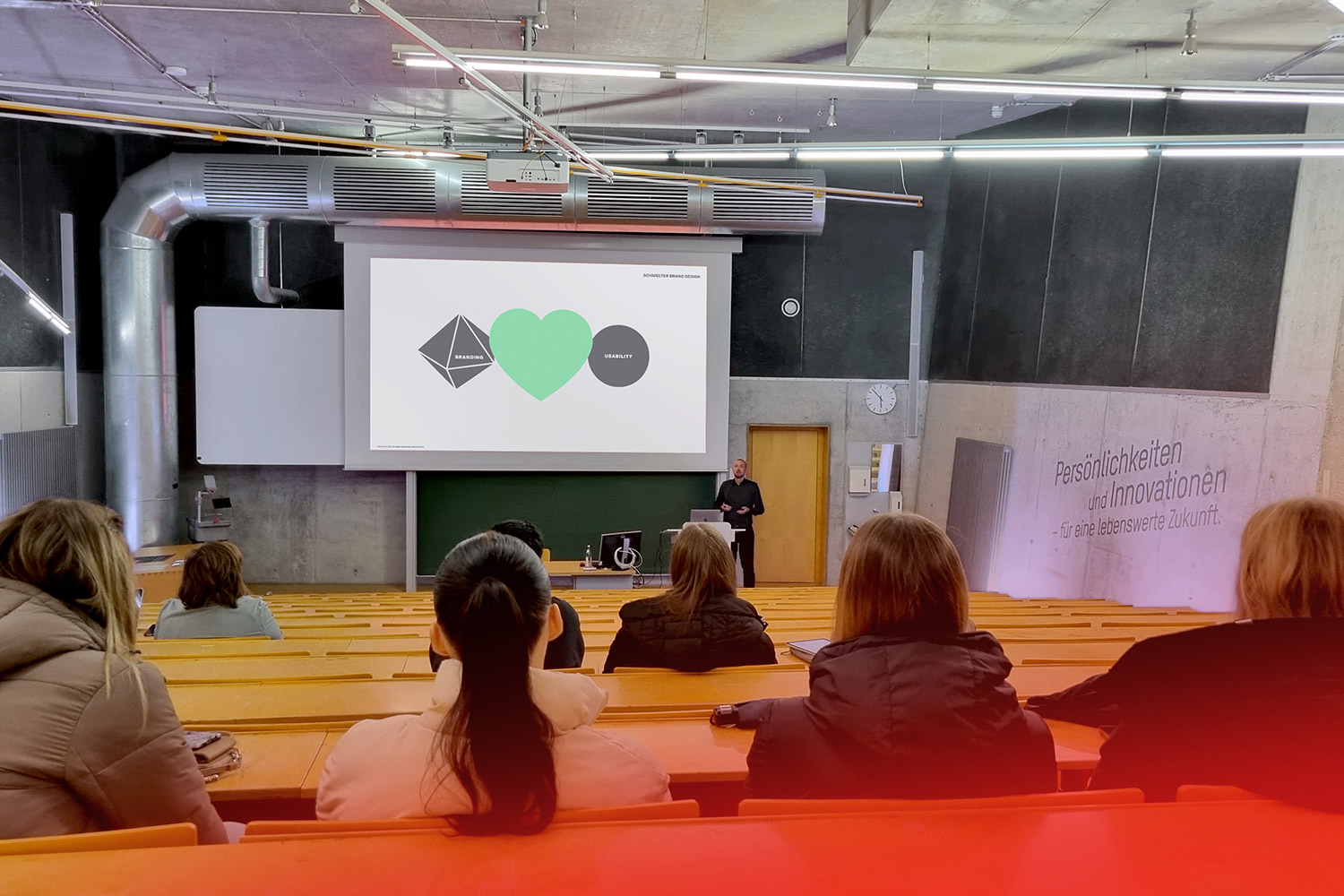 Vortrag im Hörsaal mit wenigen Zuhörern, Präsentation zeigt Symbole auf Leinwand, Fassade mit Zitat zu Persönlichkeiten.