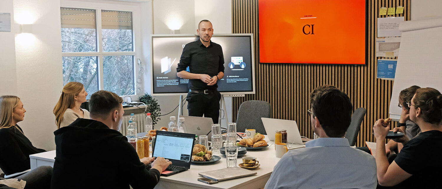Boris Schmelter hält Seminar vor Gruppe von Teilnehmern in modernem Raum mit Tisch, Laptop und Bildschirmen.