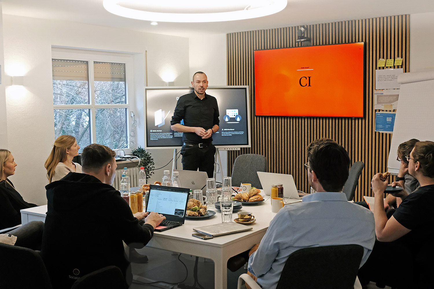 Boris Schmelter hält Seminar vor Gruppe von Teilnehmern in modernem Raum mit Tisch, Laptop und Bildschirmen.